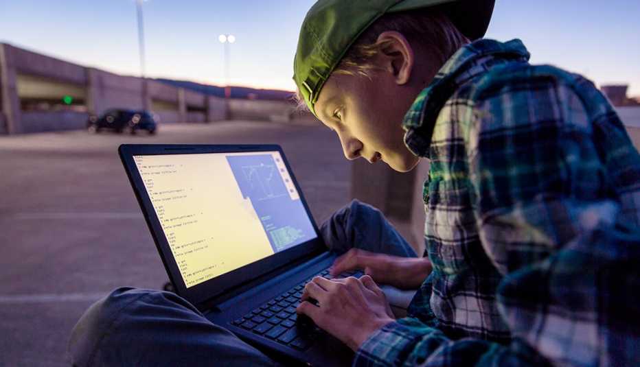 a teenage boy, who may be a scammer or being scammed, crouched over a laptop outdoors